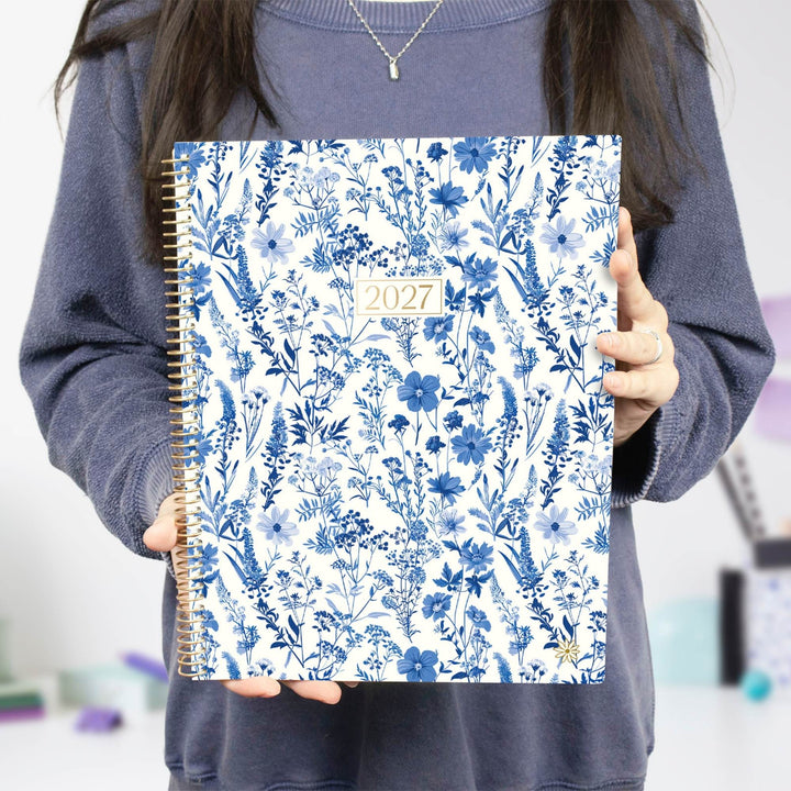 A person holding a 2027 spiral-bound planner with a blue floral pattern on a white cover, wearing a dark gray sweatshirt.
