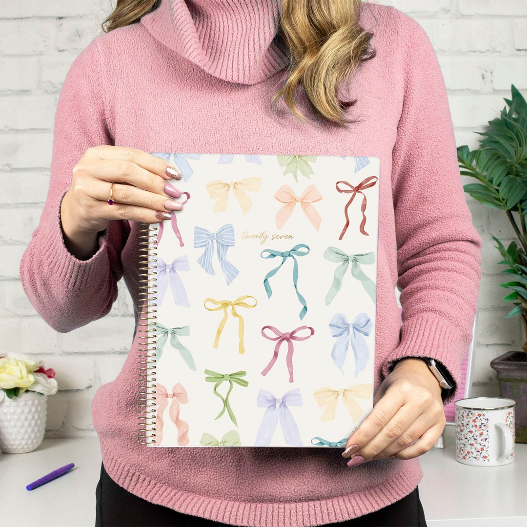 A person in a pink cowl-neck sweater holds a spiral-bound notebook with a white cover decorated with pastel ribbon illustrations. The setting is a bright, modern room with a white brick wall, a mug on a table, and a potted plant in the background.