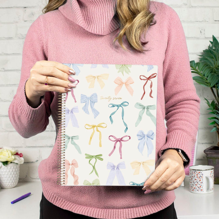 A person in a pink cowl-neck sweater holds a spiral-bound notebook with a white cover decorated with pastel ribbon illustrations. The setting is a bright, modern room with a white brick wall, a mug on a table, and a potted plant in the background.