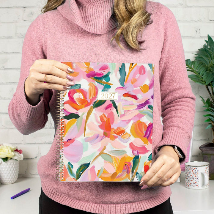 A woman in a pink turtleneck sweater holds a 2027 spiral-bound planner with a colorful floral cover. The background shows a white brick wall, a mug, and a plant.