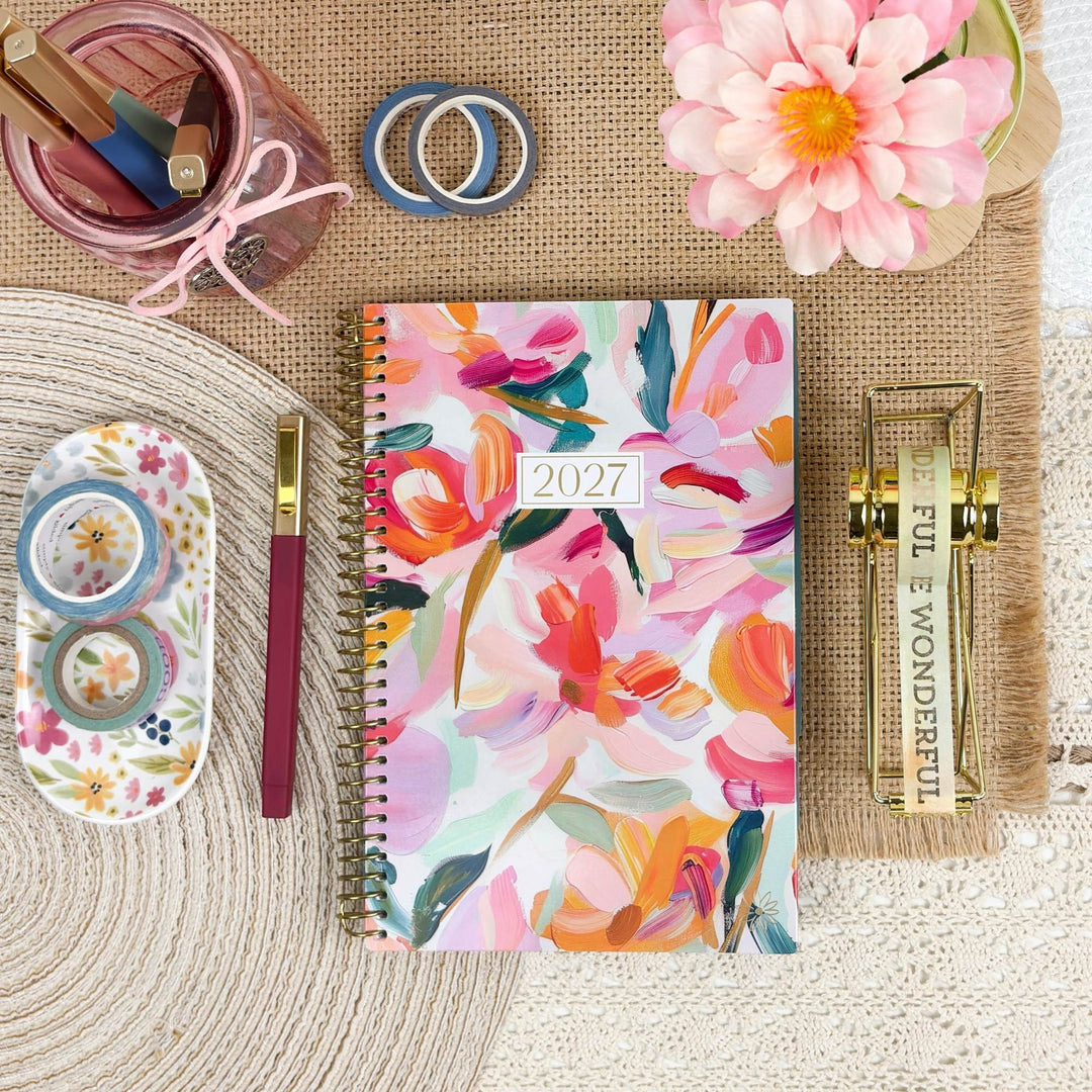 A 2027 spiral-bound planner with a colorful floral watercolor cover is arranged in a styled flat lay on a burlap mat. The planner is surrounded by desk accessories including a floral pen, washi tape, a pencil cup, and a gold journal, with a pink flower in the corner.