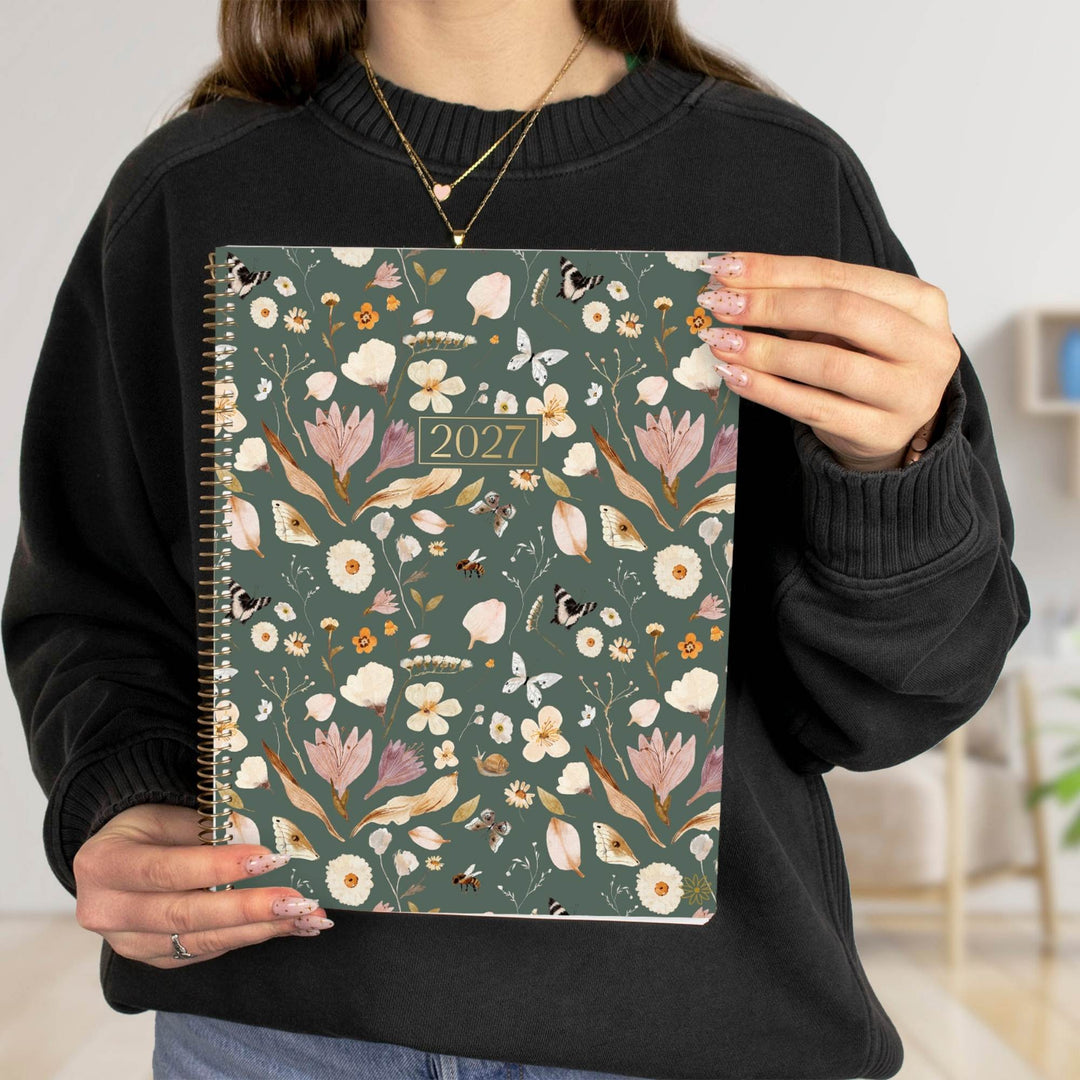 A person in a black sweatshirt holds up a 2027 spiral-bound planner with a dark green floral cover pattern featuring pink, white, and gold flowers.