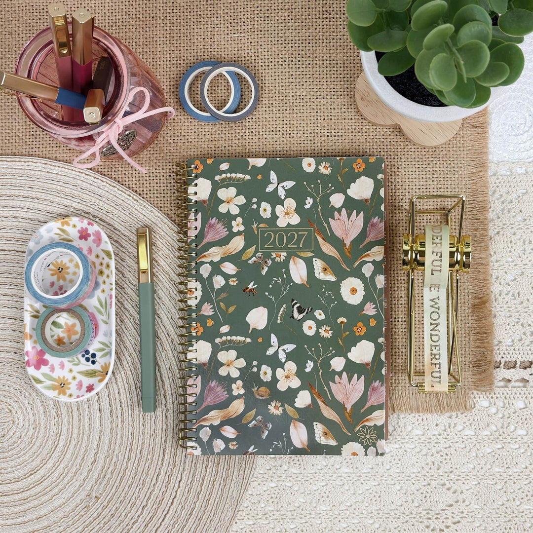 A 2027 spiral-bound planner with a green floral and bird pattern is displayed in a styled flat-lay arrangement on a burlap mat, surrounded by stationery items including a pen, washi tape, a pink cup, and a small succulent.
