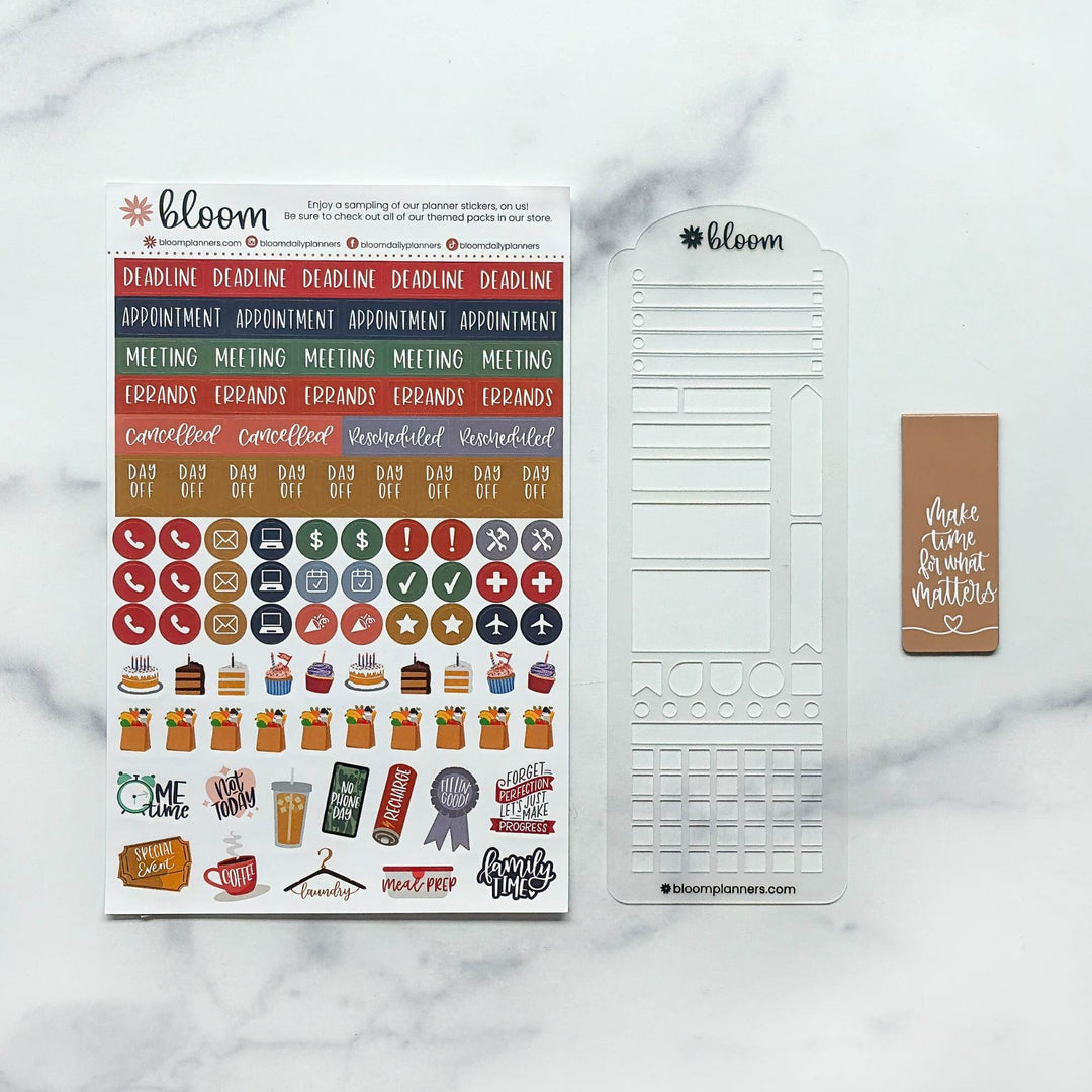 Bloom planner sticker set and clear plastic stamp sheet displayed on a white marble background. The set includes a sheet of colorful planner stickers, a clear stamp template, and a brown planner tag.
