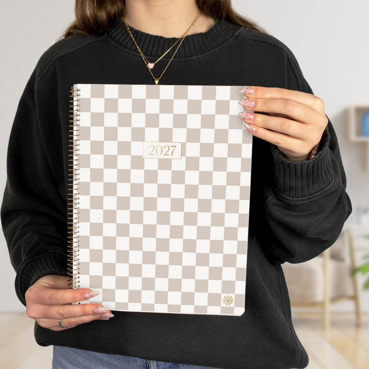 A person in a black sweatshirt holds a 2027 spiral-bound planner with a beige and white checkerboard pattern cover.