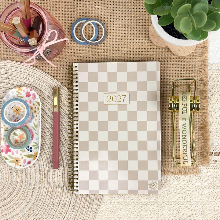 A spiral-bound 2027 planner with a beige and white checkerboard pattern, styled in a flat-lay arrangement with stationery accessories, a small succulent, and a decorative gold clip on a burlap and cream fabric background.