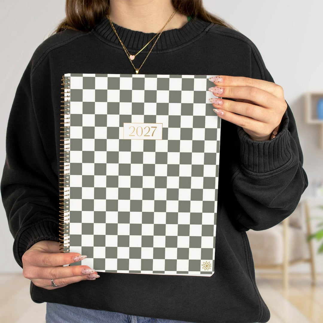 A person in a black sweater holds a 2027 spiral-bound planner with a black and white checkerboard pattern cover.