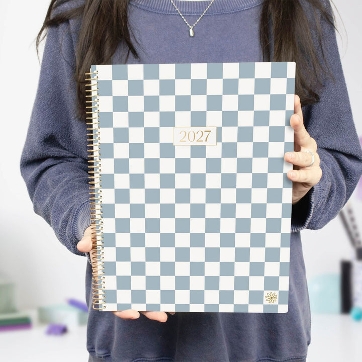 A person in a blue sweatshirt holds a 2027 spiral-bound planner with a light blue and white checkerboard pattern cover.