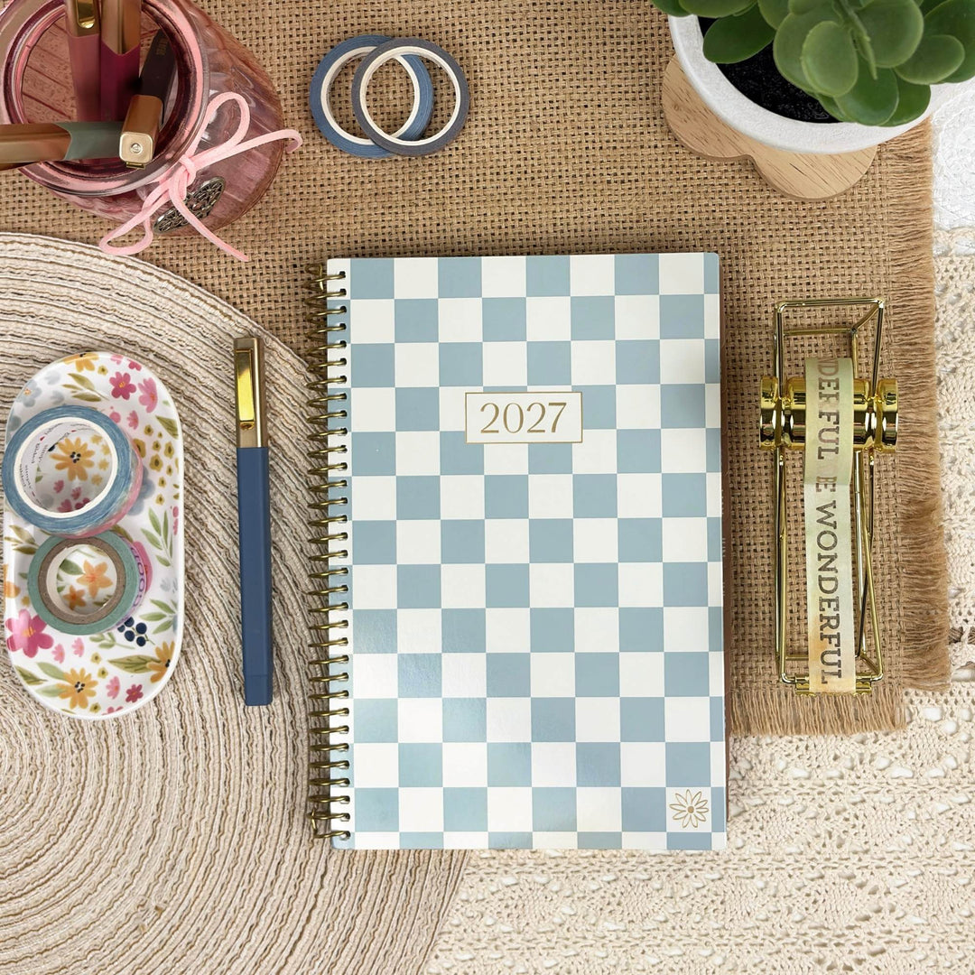 A 2027 spiral-bound planner with a light blue and white checkerboard pattern, styled in a flat-lay arrangement on a burlap mat with desk accessories.
