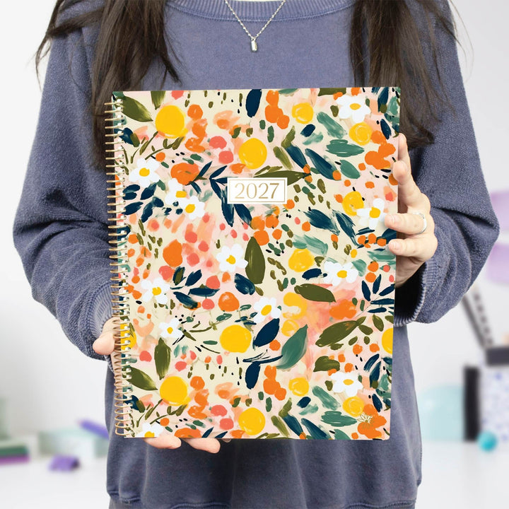 A person holding a 2027 spiral-bound planner with a colorful floral and citrus pattern on the cover, wearing a blue-gray sweatshirt.