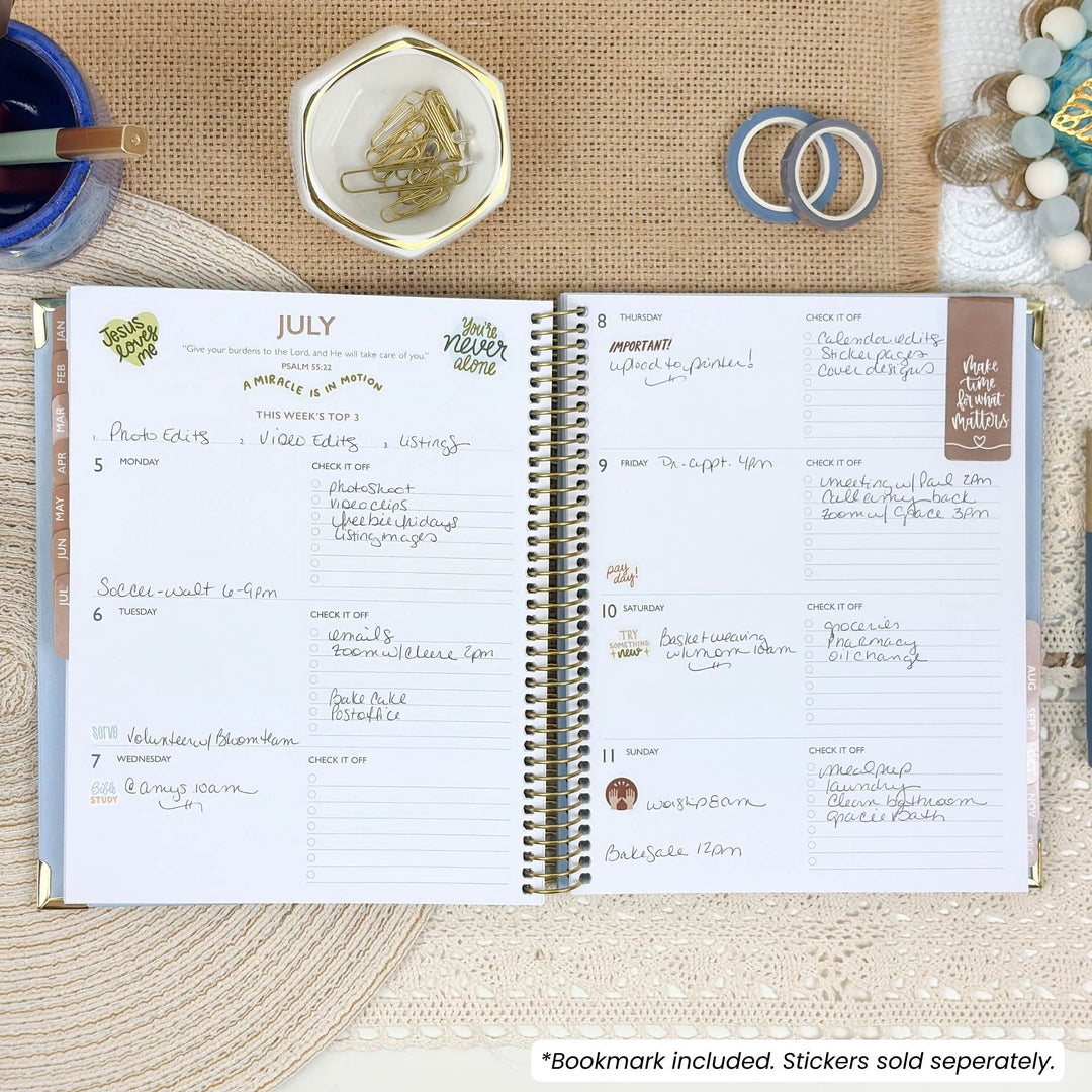 An open spiral-bound planner shown from above on a beige fabric surface, with handwritten weekly planning pages visible. Decorative props around it include a blue cup, a small dish with paper clips, two rings, and a beaded bracelet.