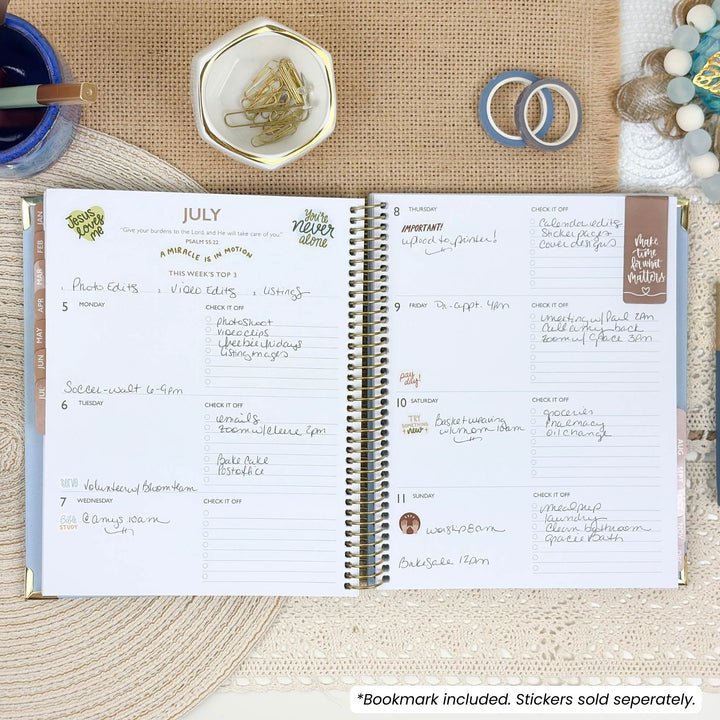 An open spiral-bound planner shown from above on a beige fabric surface, with handwritten weekly planning pages visible. Decorative props around it include a blue cup, a small dish with paper clips, two rings, and a beaded bracelet.