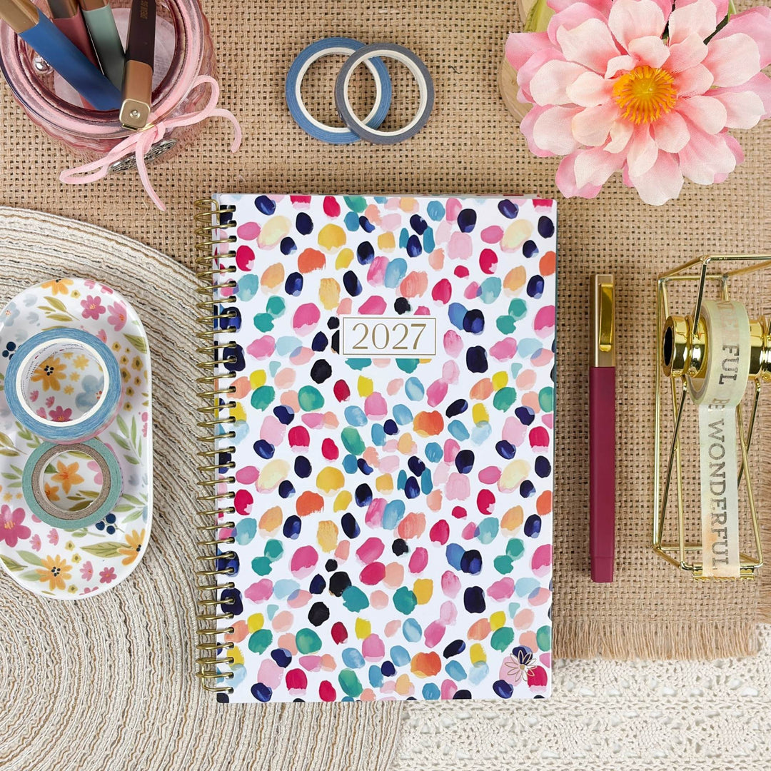 A 2027 spiral-bound planner with a colorful watercolor dot pattern on a white cover, styled in a flat-lay arrangement with stationery and decorative props on a burlap and woven mat background.