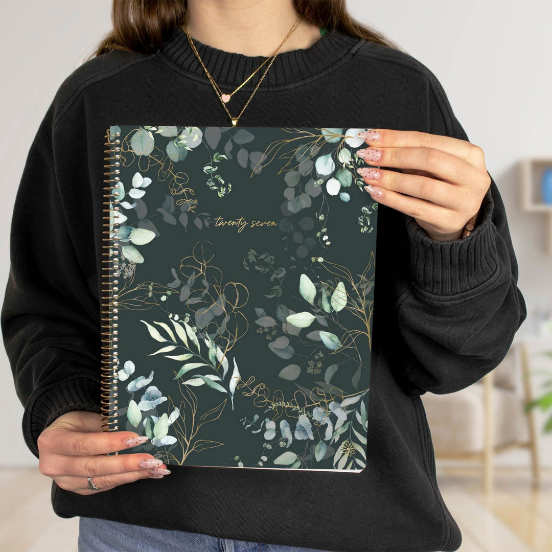 A person in a black sweatshirt holds a spiral-bound notebook with a dark green cover decorated with white and light green eucalyptus leaves and gold floral details. The cover has the text 'twenty seven' in gold script.
