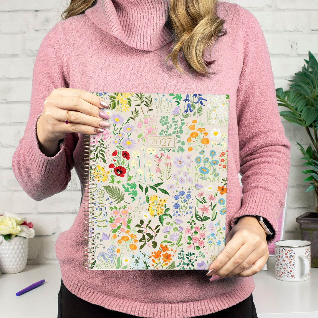 A person in a pink turtleneck sweater holds a 2027 spiral-bound planner with a colorful floral pattern. The background shows a white brick wall, a mug, and a plant.