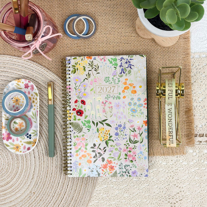 A 2027 spiral-bound planner with a colorful floral pattern is displayed in a styled flat-lay arrangement on a burlap mat, surrounded by desk accessories including washi tapes, a green pen, a small succulent, and a gold decorative object.