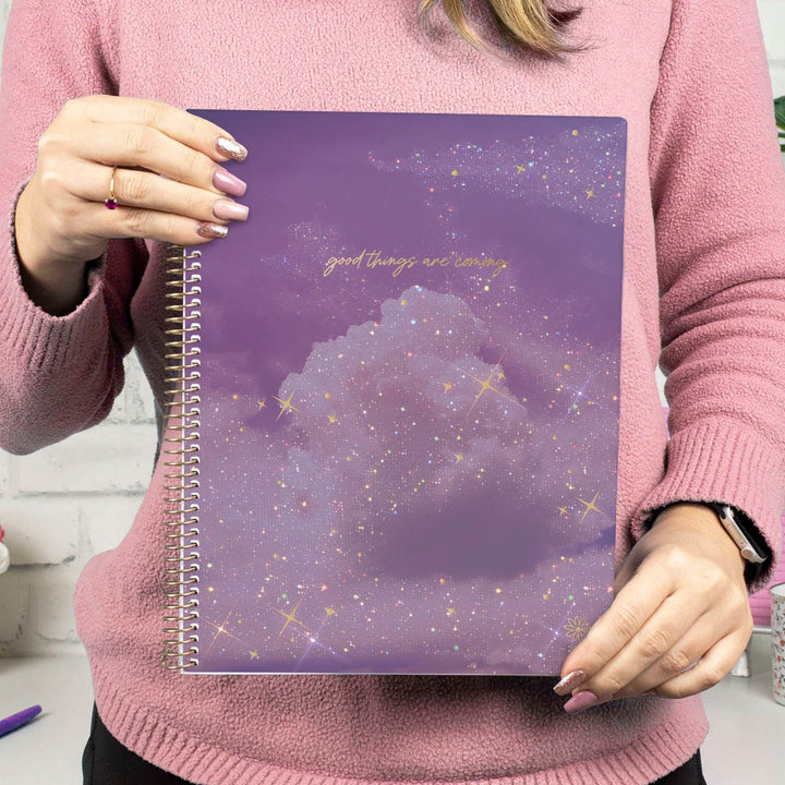 A person in a pink sweater holds a purple spiral notebook with a galaxy and cloud design, featuring the gold text 'good things are coming'.