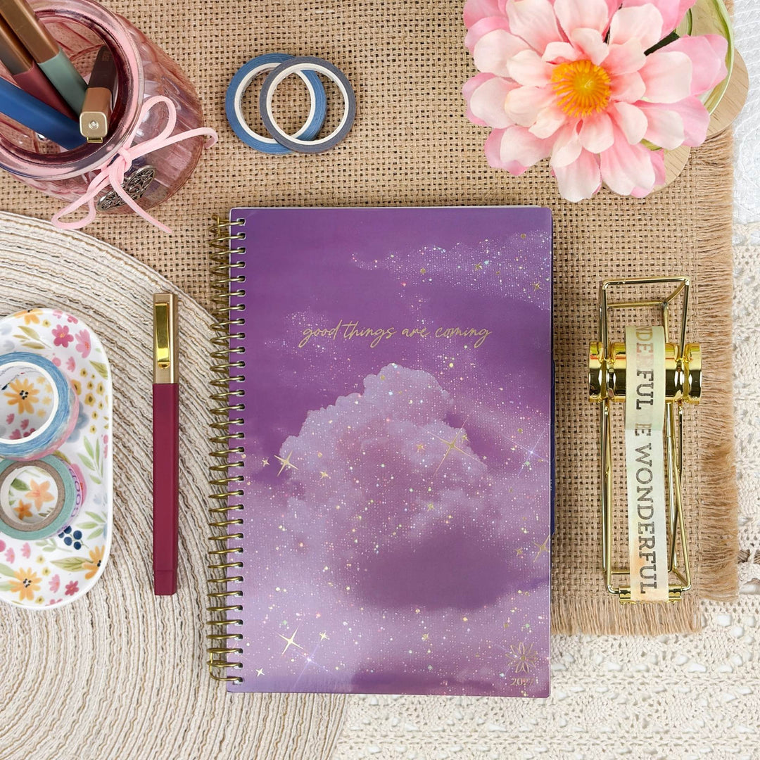 A purple spiral-bound notebook with a galaxy cloud cover design and the phrase 'good things are coming' in gold script, styled in a flat lay with stationery and decorative props on a burlap and woven surface.