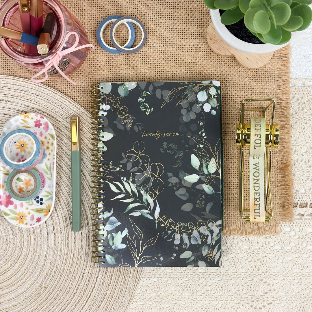 A spiral-bound planner or notebook with a dark floral cover lies on a woven mat, surrounded by desk accessories including a green pen, washi tapes, a small succulent, and a gold decorative stand.