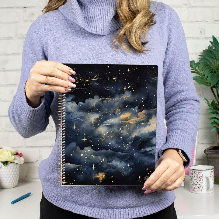 A person in a lavender turtleneck sweater holds a spiral-bound notebook featuring a dark starry sky and cloud pattern with gold glitter accents.