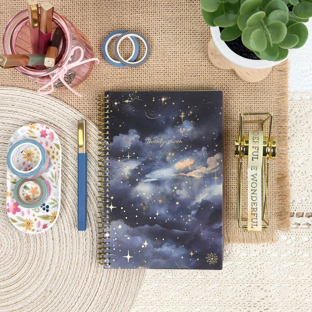 A spiral-bound notebook with a dark blue and purple starry night sky cover is arranged in a styled flat-lay on a burlap mat, surrounded by desk accessories including washi tape, a pink pencil holder, a blue pen, a small plant, and a gold decorative object.