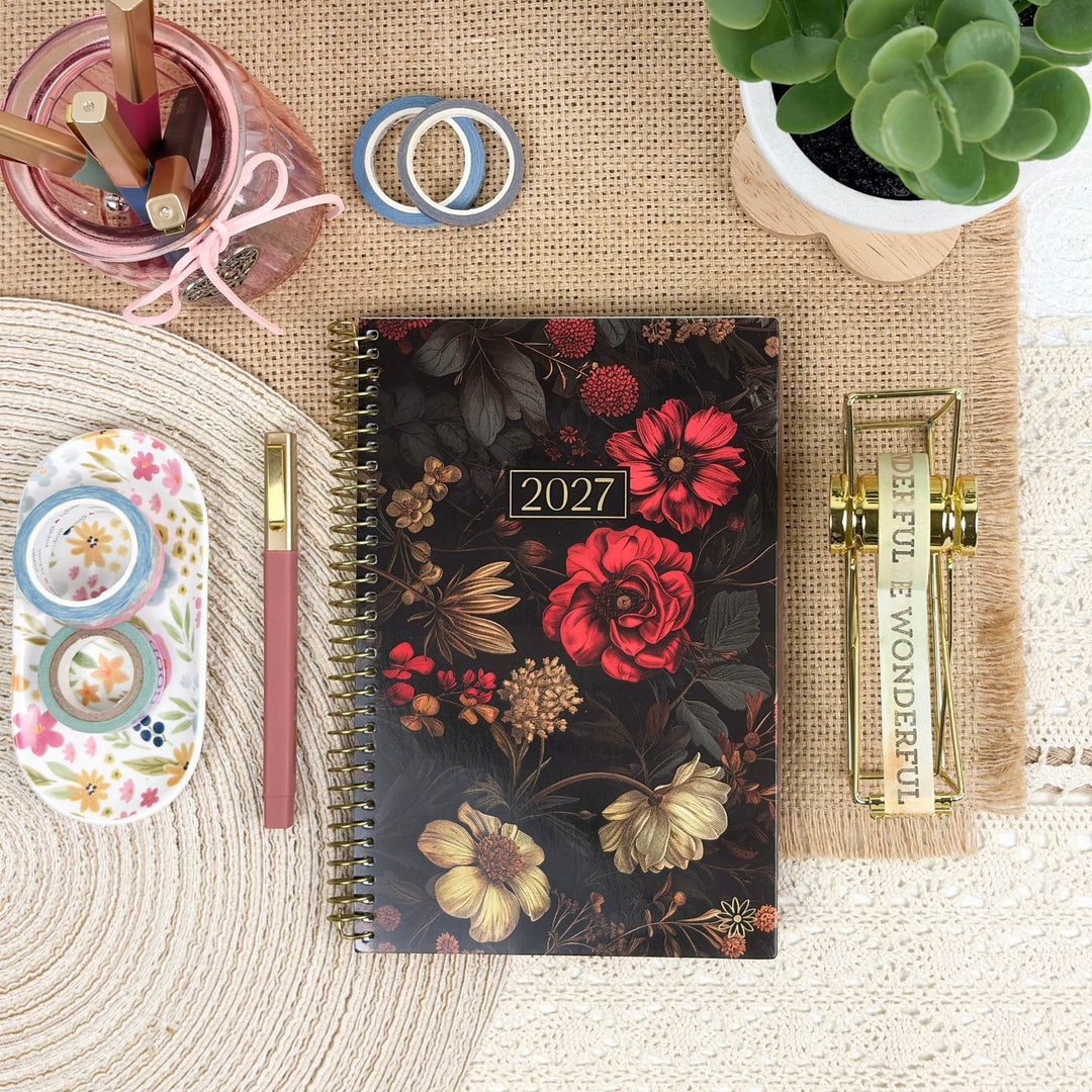 A 2027 spiral-bound planner with a dark floral cover is arranged in a styled flat lay on a burlap mat, surrounded by desk accessories including a pink cup, washi tapes, a succulent, a pen, and a gold binder clip.