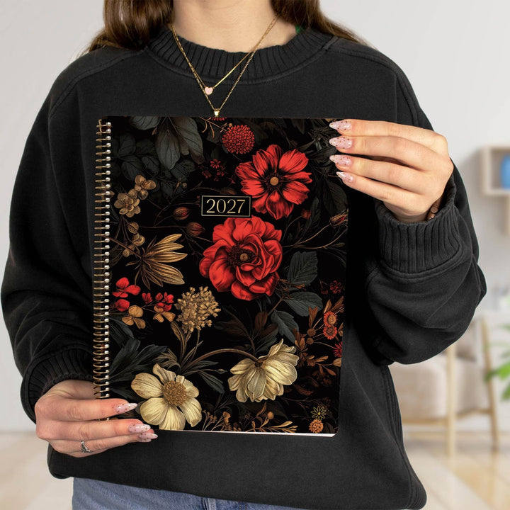 A person wearing a black sweatshirt holds a spiral-bound planner with a dark floral cover featuring red, cream, and gold flowers. The cover includes the year 2027.