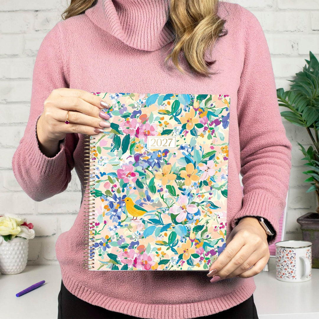 A person in a pink turtleneck sweater holds a 2027 spiral-bound planner with a colorful floral cover featuring a small yellow bird. The background shows a white brick wall, a mug, and a plant.
