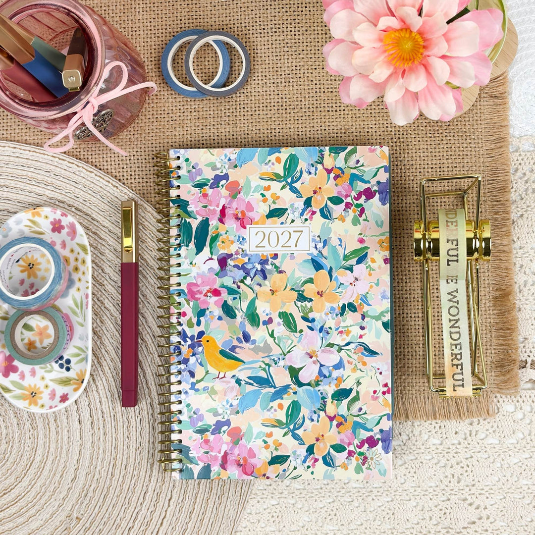 A 2027 spiral-bound planner with a colorful floral and bird pattern, styled in a flat-lay arrangement with stationery, a pink flower, and a gold letter opener on a burlap mat.