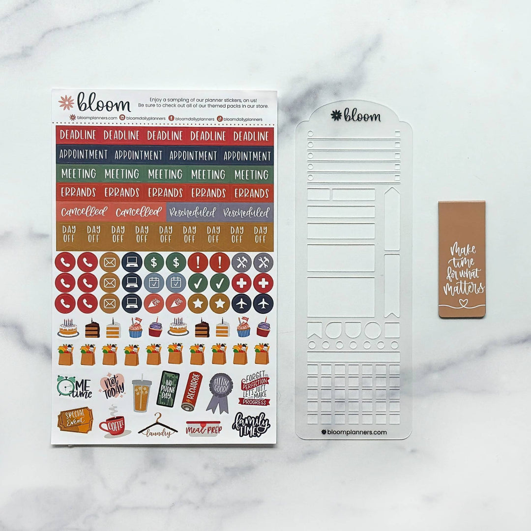 Bloom planner sticker set and clear plastic stamp sheet displayed on a white marble background. The set includes a sheet of colorful planner stickers, a clear stamp template, and a brown planner tag.