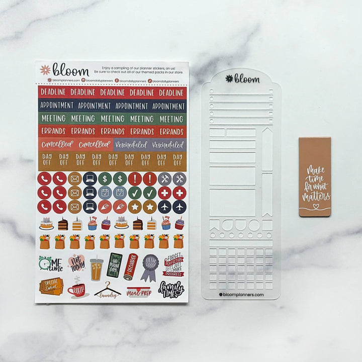 Bloom planner sticker set and clear plastic stamp sheet displayed on a white marble background. The set includes a sheet of colorful planner stickers, a clear stamp template, and a brown planner tag.