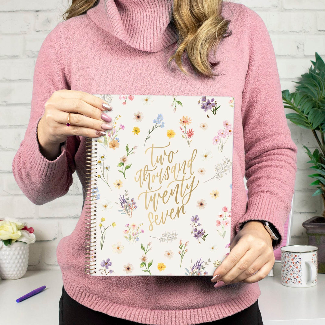 A person in a pink turtleneck sweater holds a floral spiral-bound notebook with gold script reading 'Two thousand twenty seven' against a white brick wall background.