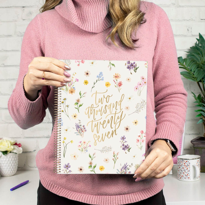 A person in a pink turtleneck sweater holds a floral spiral-bound notebook with gold script reading 'Two thousand twenty seven' against a white brick wall background.