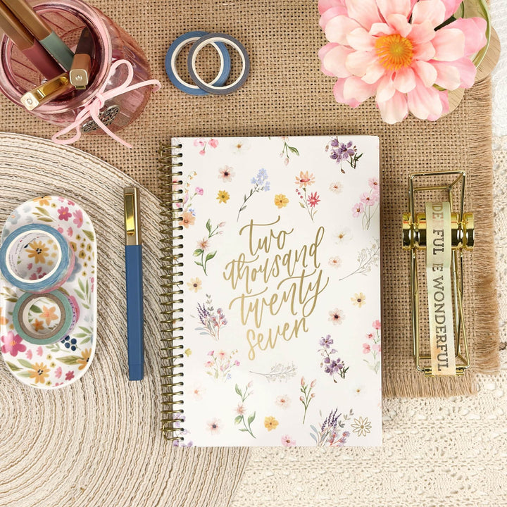 A floral spiral-bound planner or notebook with gold foil text reading 'two thousand twenty seven' is arranged in a styled flat lay on a burlap mat with decorative props including a pink flower, washi tape, a pen, and a small gold book.