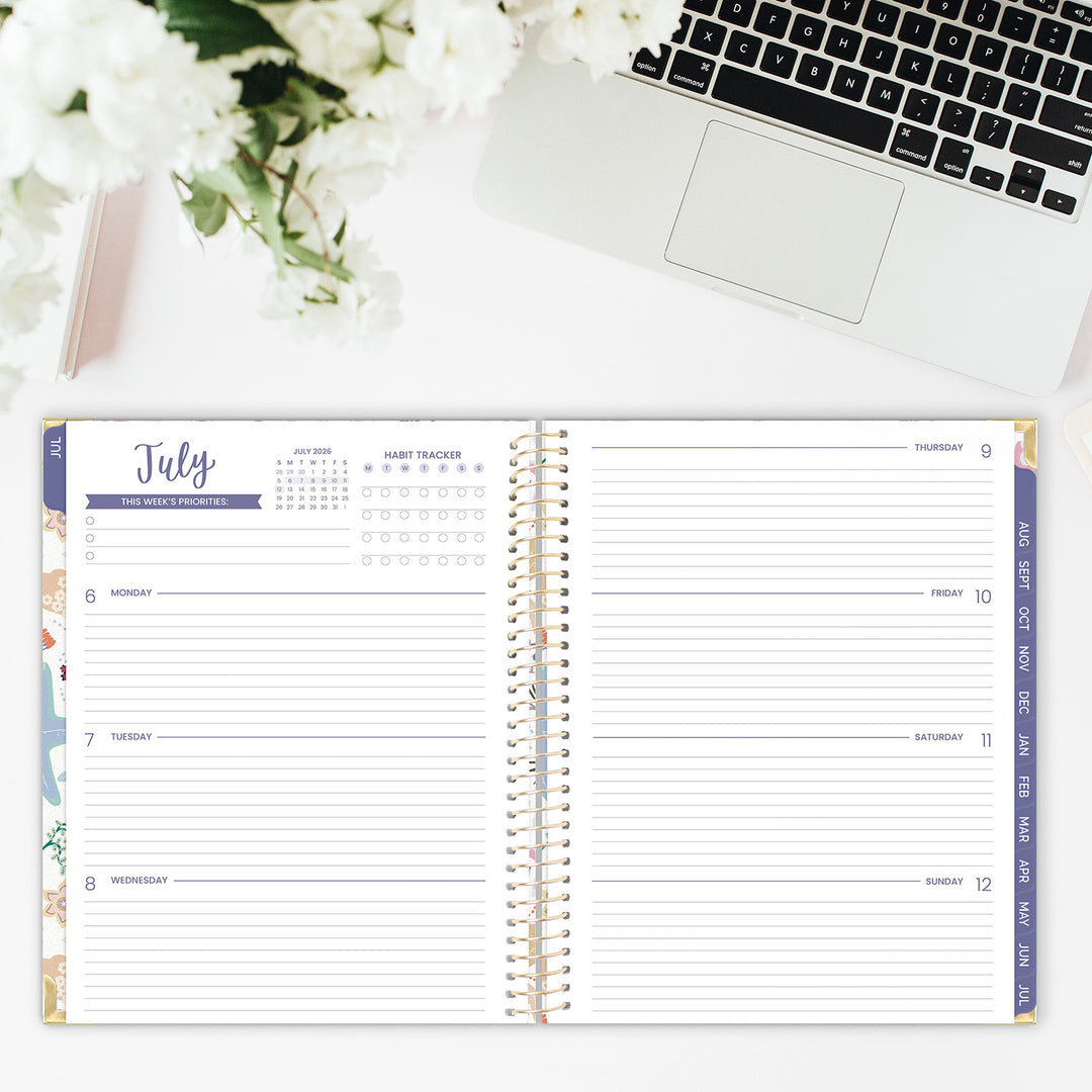 Spiral-bound planner open to a weekly spread on a desk with a laptop and flowers.