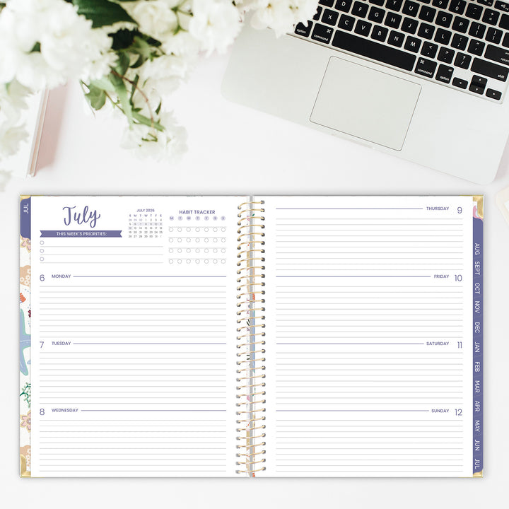 Spiral-bound planner open to a weekly spread on a desk with a laptop and flowers.