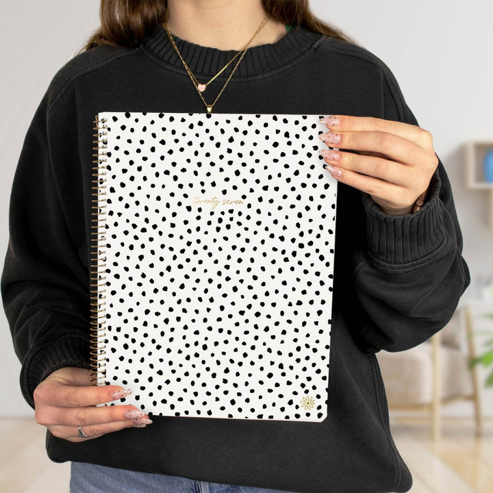A person holding a white spiral-bound planner with a black polka dot pattern and gold foil text on the cover, wearing a black sweatshirt.