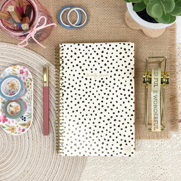 A spiral-bound planner or notebook with a white cover and black polka dots is arranged in a styled flat lay on a burlap mat, surrounded by stationery items including washi tapes, a pink pen, a floral tray, a pencil cup, and a small plant.