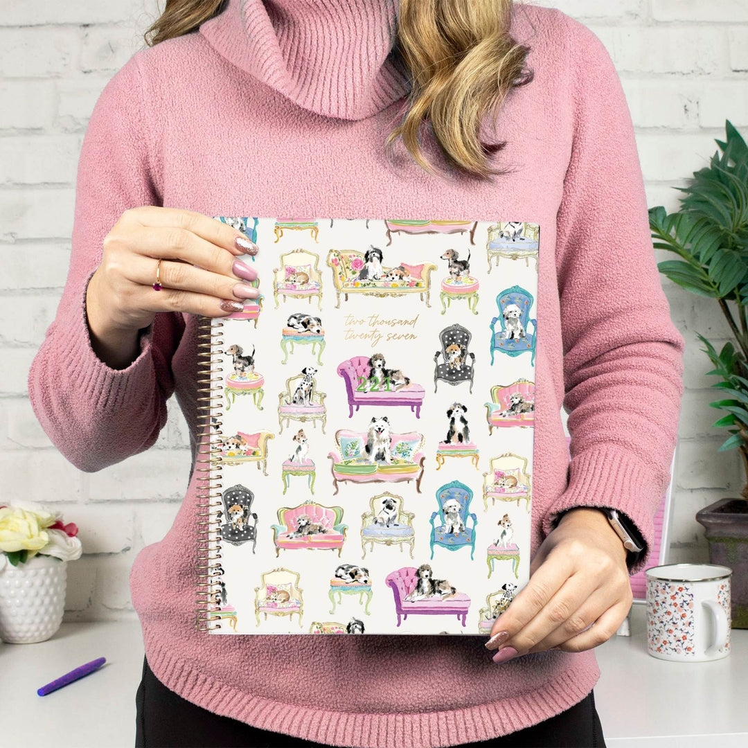 A person in a pink turtleneck sweater holds a spiral-bound notebook featuring a whimsical pattern of dogs lounging in colorful armchairs. The background shows a white brick wall, a plant, a mug, and a small flower pot.