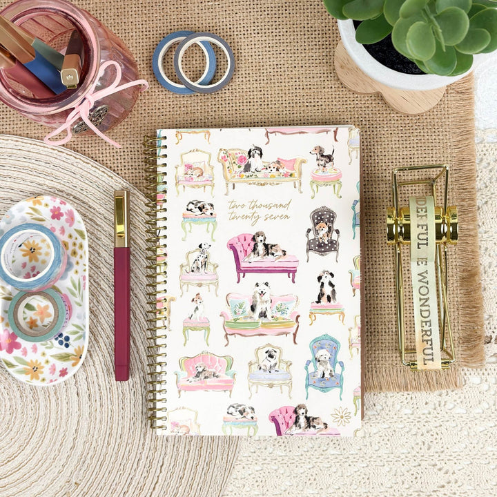 A spiral-bound planner or notebook with a white cover decorated with pastel illustrations of dogs lounging in pink and purple chairs, styled in a flat lay on a burlap mat with stationery accessories around it.