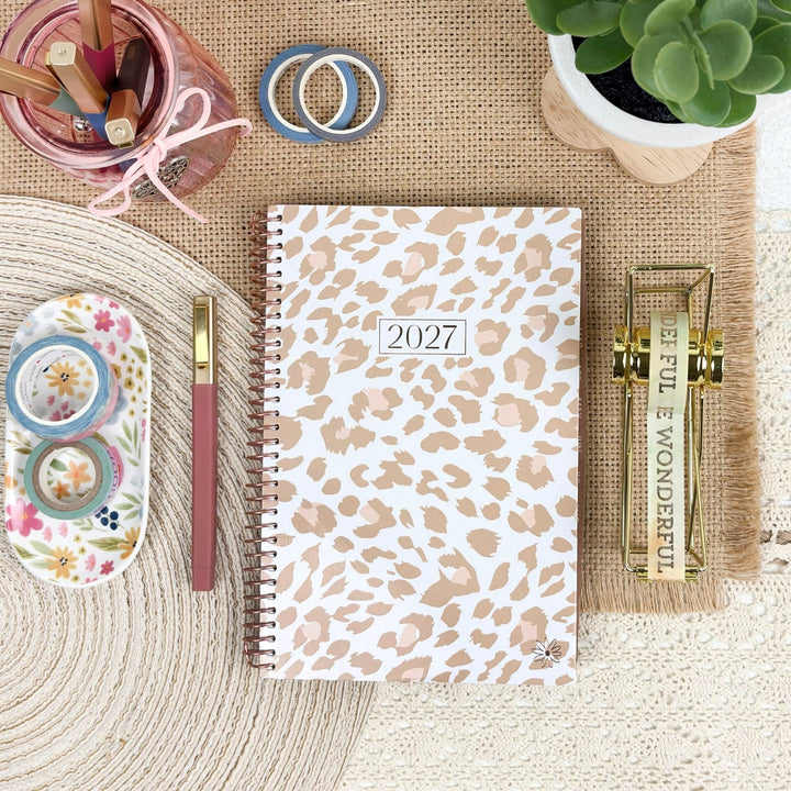 A 2027 spiral-bound planner with a beige and white leopard print cover is displayed in a styled flat-lay arrangement on a burlap mat. The planner is surrounded by desk accessories including a pink pen, washi tapes, a floral dish with rolls, a pink pencil cup, and a small potted succulent.