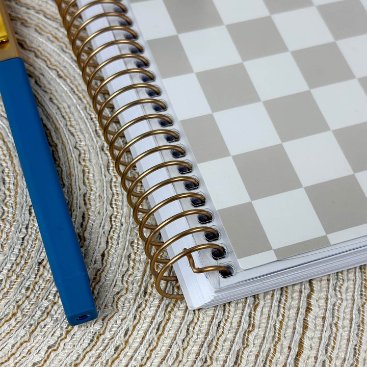 A spiral-bound notebook with a white and gray checkerboard cover, placed on a textured beige woven mat next to a blue pen.