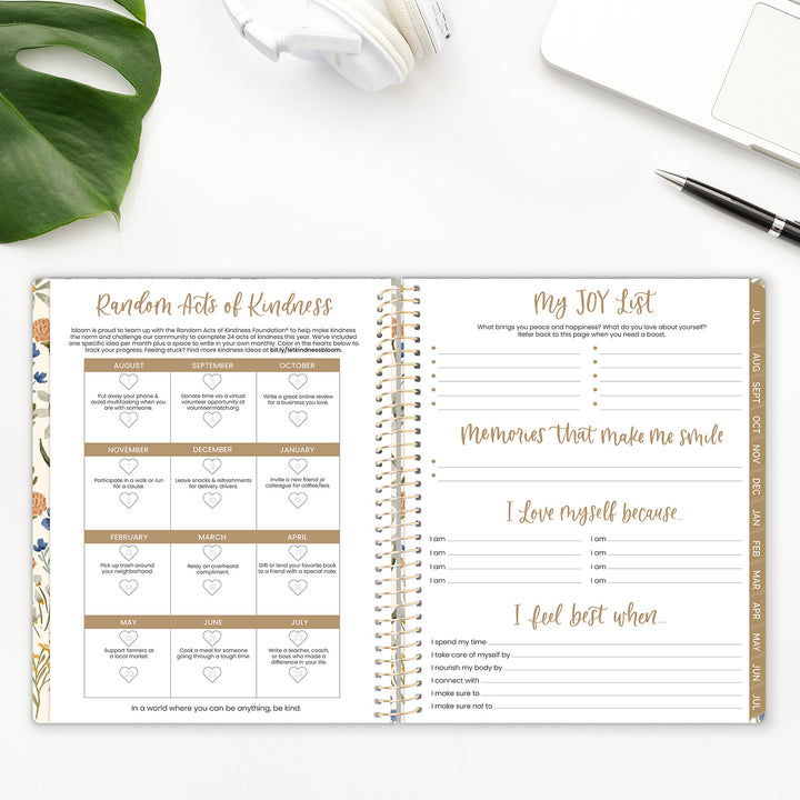 Open planner with sections for Random Acts of Kindness and a JOY List on a white surface with a plant and pen.