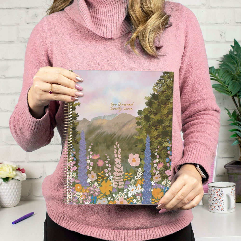 A woman in a pink turtleneck sweater holds a spiral-bound planner or notebook featuring a colorful landscape illustration of mountains, trees, and wildflowers. The text on the cover reads 'Our Town Beautiful Town. - hover