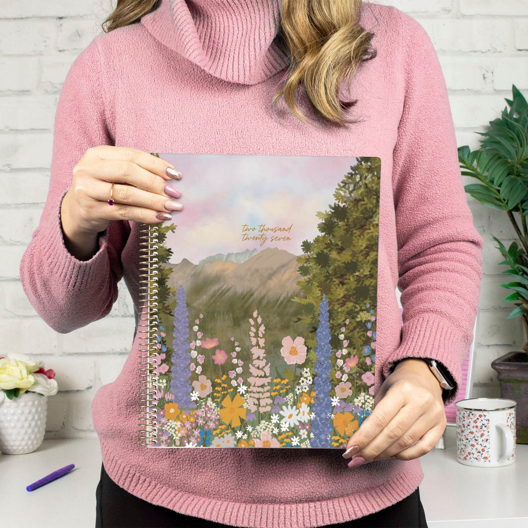 A woman in a pink turtleneck sweater holds a spiral-bound planner or notebook featuring a colorful landscape illustration of mountains, trees, and wildflowers. The text on the cover reads 'Our Town Beautiful Town.