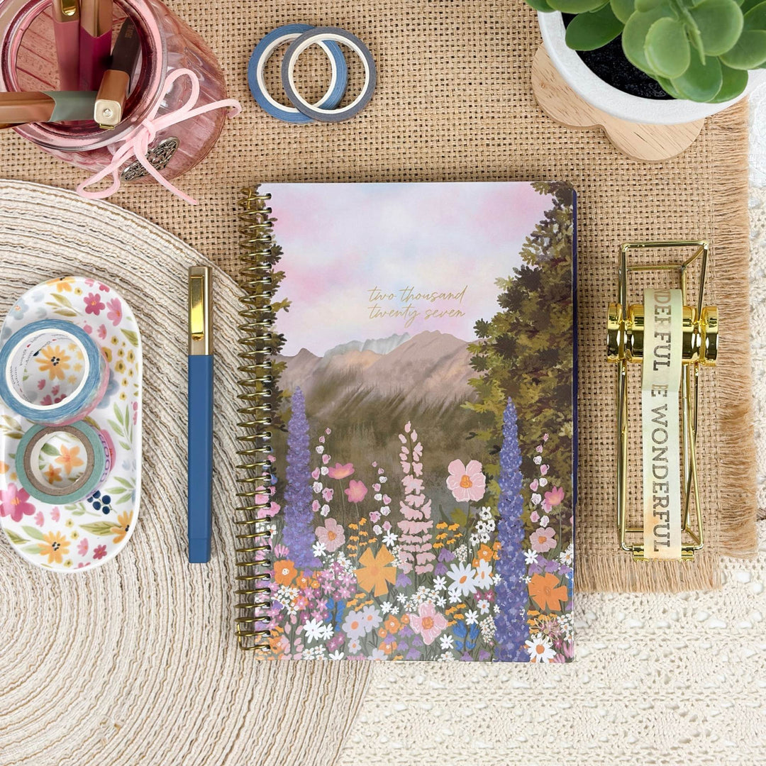 A spiral-bound planner or notebook with a floral mountain landscape cover is arranged on a woven mat with stationery props, including washi tape, a blue pen, a small plant, and a gold clip.