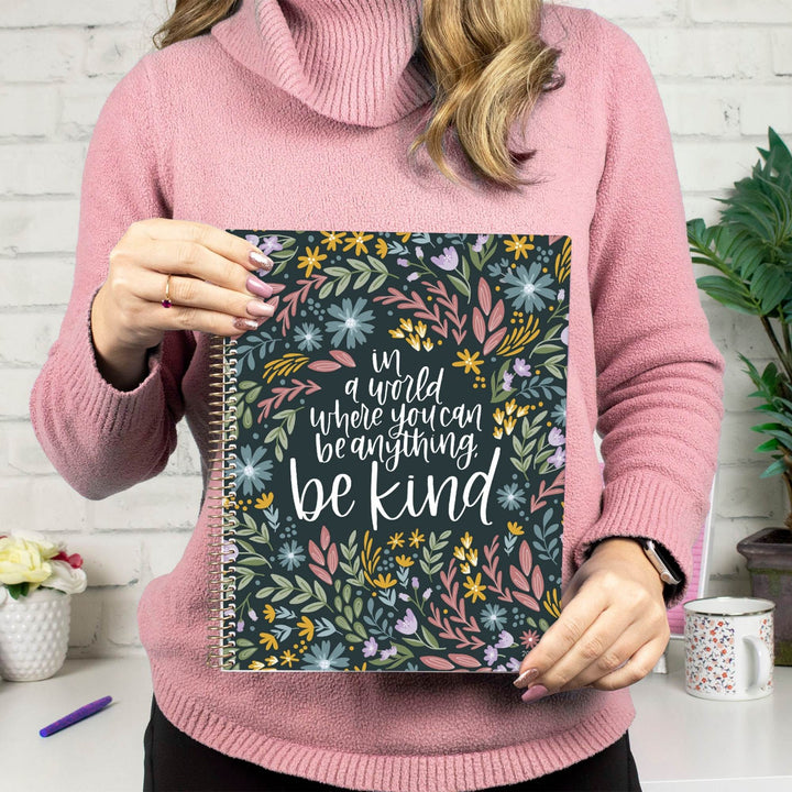 A person in a pink turtleneck sweater holds a floral notebook or planner cover with the phrase 'in a world where you can be anything be kind' in white script. The cover has a dark green background with colorful flowers in yellow, pink, blue, and purple.
