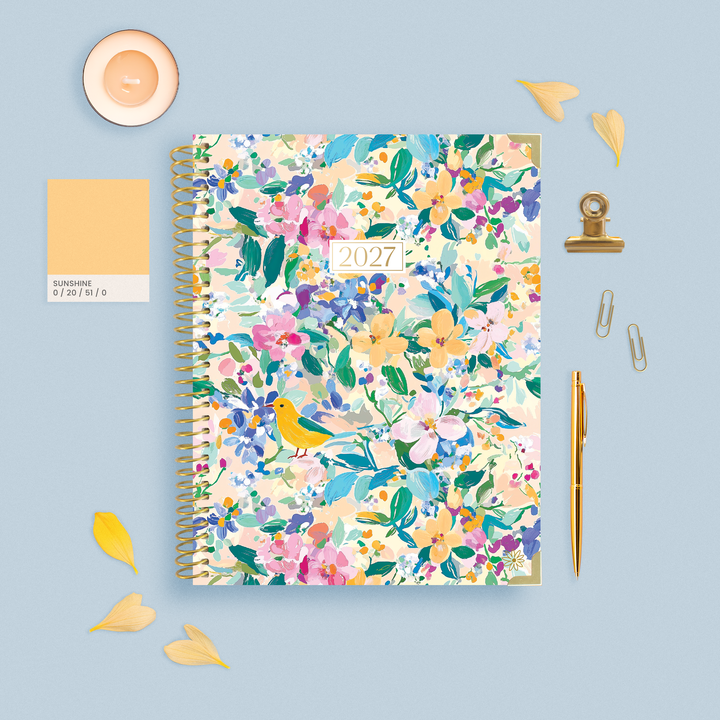 A 2027 spiral-bound planner with a colorful floral and bird pattern, styled in a flat lay on a light blue background with a candle, color swatch card, clips, pen, and paper clips.