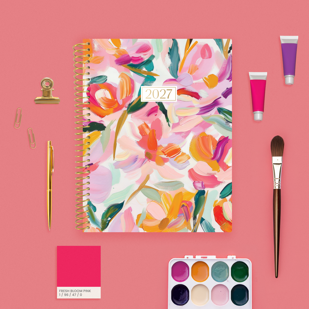 A 2027 spiral-bound planner with a colorful floral watercolor cover is arranged on a pink background with stationery and art supplies, including a gold pen, binder clip, paintbrush, paint tubes, a small paint palette, and a pink color card.