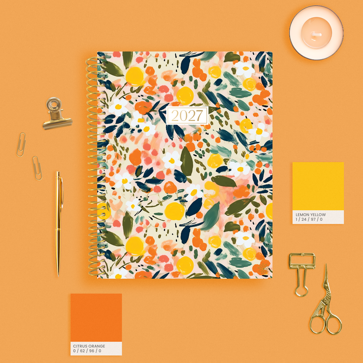 A 2027 spiral-bound planner with a colorful watercolor floral and citrus pattern, styled in a flat-lay on an orange background with gold stationery and color swatch cards.